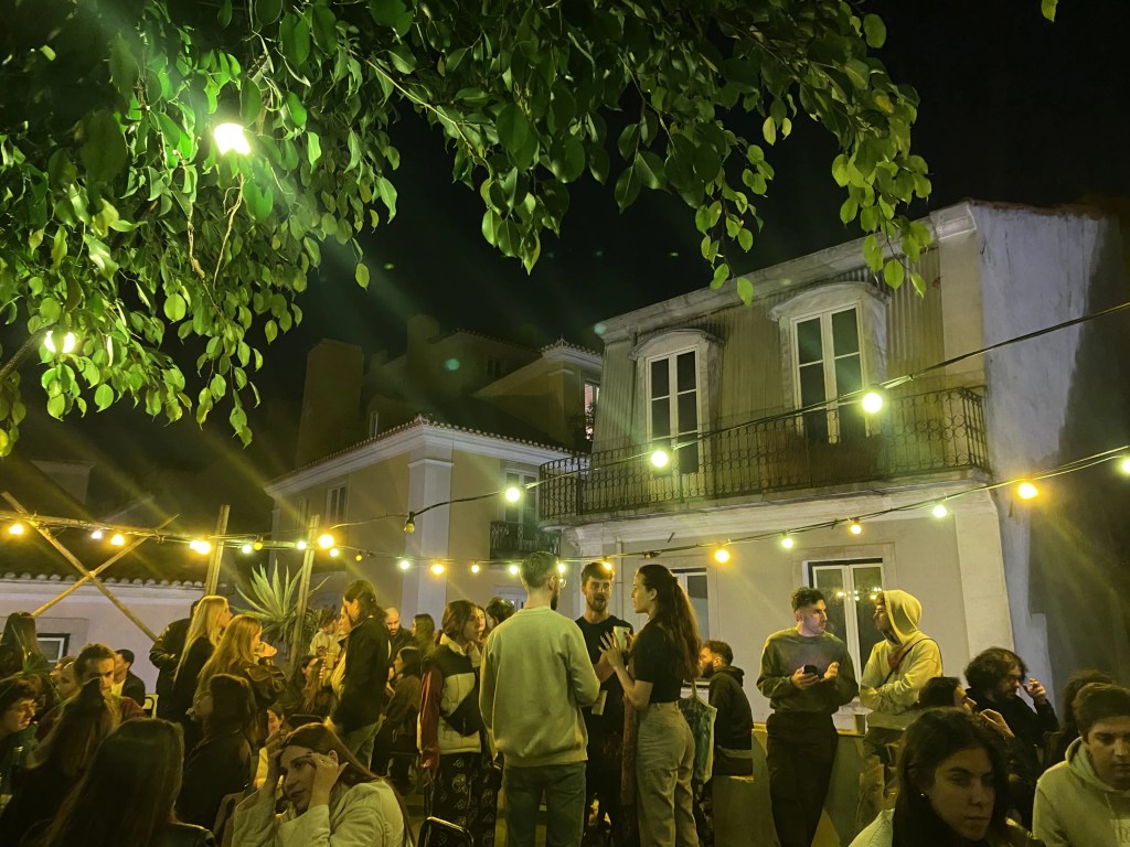 A lively outdoor gathering at night in Lisbon, Portugal with people socializing under string lights, surrounded by trees and a building in the background. After a surf lesson in Costa da Caparica, socialising with your friends sounds like the think to do!