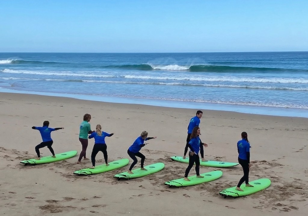 A group of six surfers in a surf lesson practicing on surfboards on the beach at Breakthrough Surf School in Costa da Caparica near Lisbon, Portugal